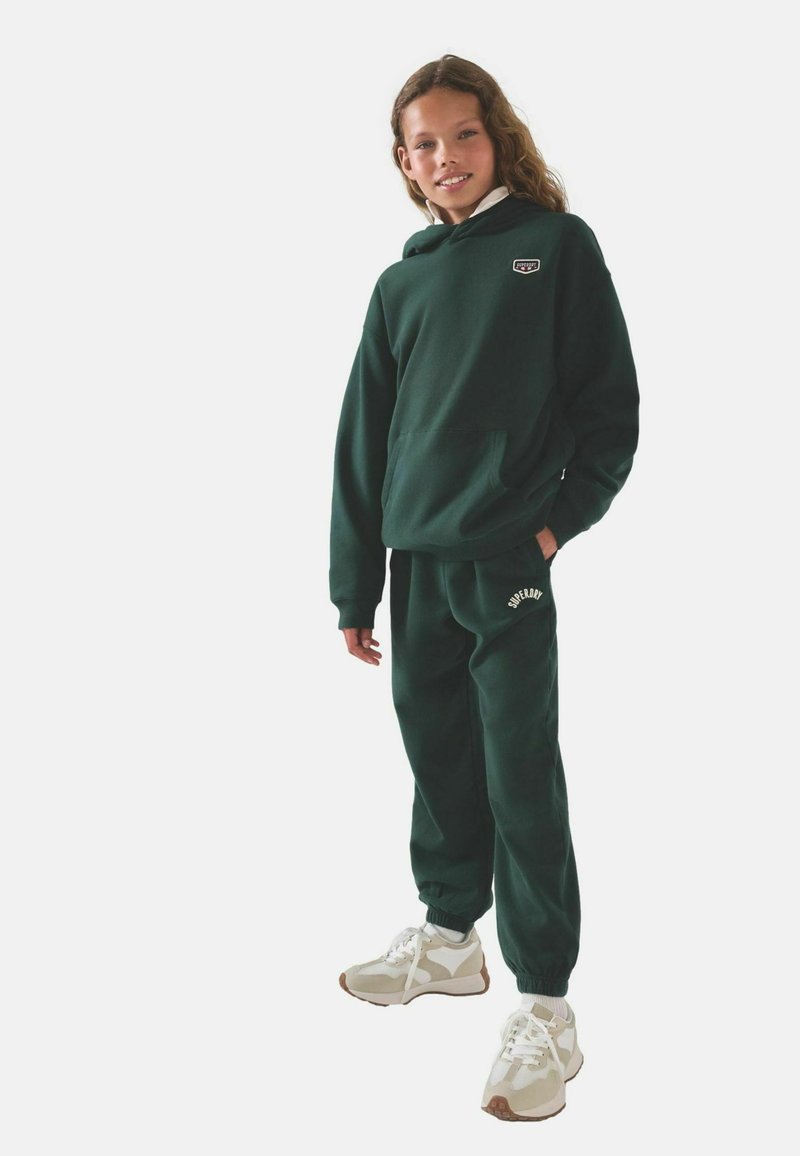 Felpa con cappuccio verde in cotone con tasca frontale, abbinata a pantaloni della tuta coordinati. Presenta loghi sulla felpa e sui pantaloni, completati da sneakers bianche.