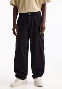 Pantaloni cargo neri con una vestibilità rilassata, dotati di tasche applicate e dettagli di cucitura a vista. Abbinati a sneaker bianche e una camicia beige.