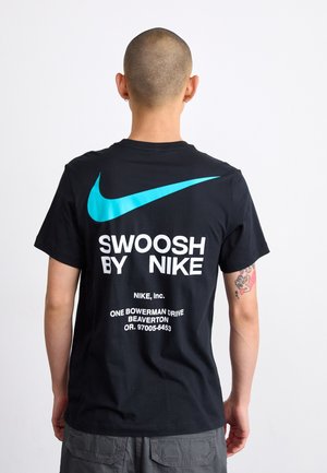 Person med barberet hoved iført sort Nike T-shirt med stor blå swoosh og hvid tekst på bagsiden, stående mod en ensfarvet baggrund.
