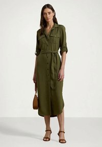 Γυναίκα σε ελαιόχρωμο μεσάτο φόρεμα με κουμπιά και γυρισμένα μανίκια, με ζώνη στη μέση, κρατώντας καφέ τσάντα και φορώντας καφέ πέδιλα με τακούνι.