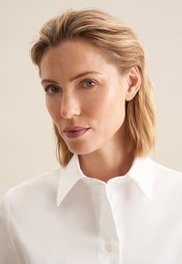 Hvid skjorte med krave lavet af glat stof, med knaplukning og en figursyet design. Rent, minimalistisk stil uden synlige mønstre.