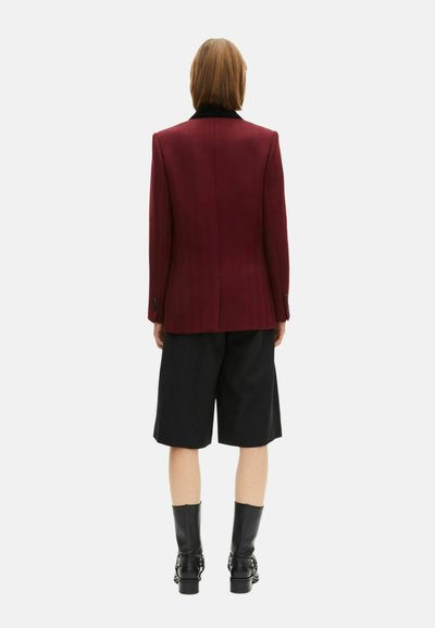 Blazer bordeaux con colletto nero, abbinato a pantaloni corti neri. Il blazer presenta un design strutturato e una vestibilità sartoriale, realizzato in tessuto testurizzato.