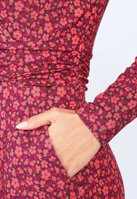 Figurbetontes Kleid aus lila Stoff mit einem floralen Druck in Pink, grünen Akzenten, langen Ärmeln und einer seitlichen Tasche. Strukturierter Stoff.