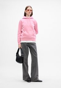 Sudadera con capucha rosa y bolsillo frontal, encima de una camisa blanca. Combinada con pantalones grises de pierna ancha y un bolso negro con correa.