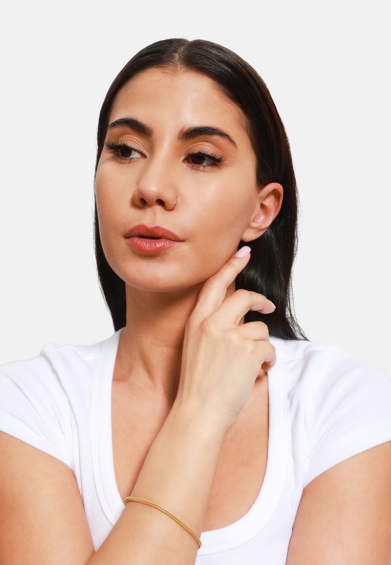Gold bracelet with a textured design, worn on the wrist. Subject with dark hair poses, wearing a white fitted top.