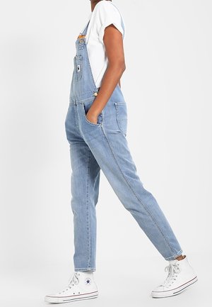 Osoba ubrana w jasnoniebieskie ogrodniczki z denimu i białe wysokie trampki Converse, stojąca z rękami w kieszeniach na jednolitym tle.