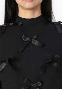 Top negro texturizado que presenta múltiples pequeños lazos de satén negro como embellishments. El escote es alto y la tela está ligeramente arrugada.