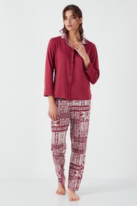 Kvinde iført vinrød langærmet bluse med knapper og mønstrede vinrøde og hvide pyjamabukser, stående barfodet mod hvid baggrund.