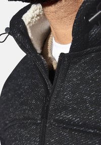 Black textured jacket featuring a zip closure, lined with cream fleece around the collar, and layered over a light-colored shirt.