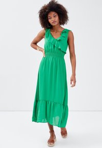 Robe maxi verte avec un col à volants et une taille smockée. Présente une jupe à volants et un tissu léger. Portée avec des sandales espadrilles marron.