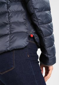 Primo piano di una persona che indossa un giubbotto trapuntato blu navy con una cerniera rossa e jeans di denim blu scuro.