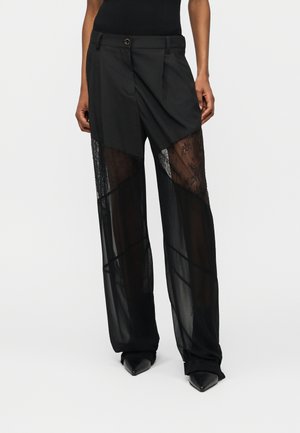 Žena v čiernych nohaviciach s širšími nohavicami so sieťovanými a priesvitnými panelmi, v čiernych špicatých topánkach a v čiernom tielku bez rukávov, stojaca na bielom pozadí.