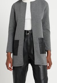 Nainen, jolla on yllään teksturoitu harmaa avomallinen neule, jossa on mustat nahkapaneelit taskuissa, valkoinen ribbattu poolopaita ja mustat vyölliset nahkahousut.