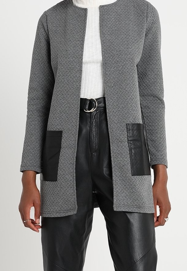 Nainen, jolla on yllään teksturoitu harmaa avomallinen neule, jossa on mustat nahkapaneelit taskuissa, valkoinen ribbattu poolopaita ja mustat vyölliset nahkahousut.