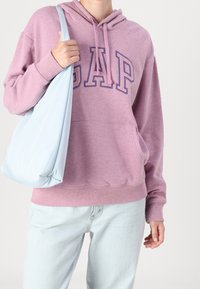 Różowa bawełniana bluza z kapturem z napisem "GAP" w ciemniejszym odcieniu fioletu. W komplecie z jasnoniebieską torbą na ramię i jasnymi dżinsami. Posiada kieszeń z przodu i kaptur.