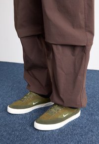 Zelené semišové a textilní tenisky s bílým logem Nike, spárované s volnými hnědými kalhotami, na texturovaném modrém koberci.