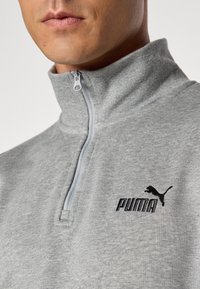 Šedá mikina na zip s vysokým límcem, která má jemnou texturu a černé logo Puma na levém prsníku. Zip má bílou kontrastní úchytku.