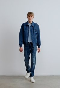 Ναυτικό μπουφάν bomber με λεία υφή, ανοιχτό μπλε πουκάμισο από κάτω, φορεμένο με μπλε τζιν και λευκά αθλητικά παπούτσια, στέκεται μπροστά σε φωτεινό φόντο.