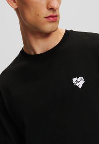 Pulover din bumbac negru, cu guler rotund și un mic logo brodat în formă de inimă albă în partea stângă a pieptului.