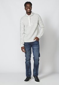 Hellgrauer gestrickter Kapuzenpullover mit einem Schalkragen, weißem Kordelzug; kombiniert mit dunkelblauen Jeans und schwarzen Turnschuhen. Glatte Textur, lässige Passform.