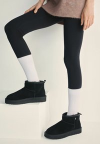 Czarne zamszowe botki do kostki na płaskiej podeszwie z pętelką z tyłu, w połączeniu z czarnymi legginsami i białymi skarpetkami w prążki.