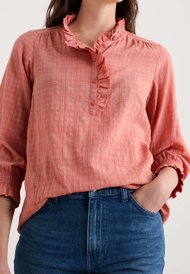 Femme portant un chemisier corail à volants avec boutons et manches trois-quarts, rentrée dans un jean bleu taille haute, main dans la poche.