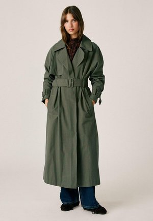 Joven mujer que lleva un largo trench verde con cinturón, top oscuro con estampado, vaqueros azules y zapatos negros, de pie con las manos en los bolsillos del abrigo.