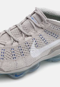 Gray knitted athletic shoe with blue accents, transparent sole, and white swoosh logo. Features a breathable texture and elastic lacing system.
