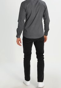 Czarna koszula w wzorki z długim rękawem, zestawiona z czarnymi spodniami i białymi trampkami. Koszula ma mały geometryczny nadruk.