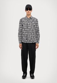 Black and white long-sleeve shirt with bold text pattern, paired with loose black pants and black sneakers, complemented by dark sunglasses and cap.
