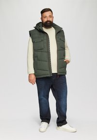 Grüne Steppweste mit Kapuze, Fronttaschen und Reißverschluss über einem cremfarbenen Pullover. Getragen mit dunklen Jeans und weißen Sneakers.