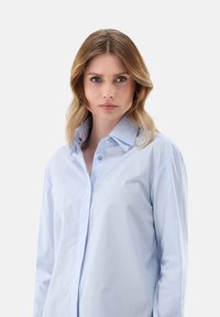 Donna con capelli biondi fino alle spalle, indossa una camicia azzurro chiaro con bottoni, rivolta in avanti con un'espressione neutra su uno sfondo semplice.