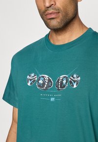 T-shirt de algodão em teal com uma estampa gráfica de três anéis e o texto "HISTORY MAKERS CITY" em branco, com destaques em preto e azul.