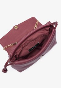 Sac à main en cuir bordeaux avec une bandoulière en chaîne dorée, fermeture à pression et intérieur en tissu portant une étiquette "ANNA FIELD".