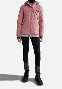 Světle růžová prošívaná bunda s kapucí, černými akcenty a logem. Nosená přes kostkovanou košili, v kombinaci s černými legínami a černými botami na tkaničky.