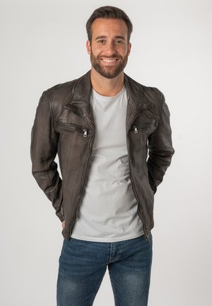 Braune Leder-Motorradjacke mit Reißverschlusstaschen und strukturierter Oberfläche, kombiniert mit einem hellgrauen T-Shirt und blauen Jeans.