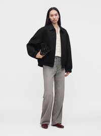 Femme debout portant une veste noire, un pull en maille blanc, un jean gris à jambes larges, des chaussures bordeaux, et tenant un sac à main noir sur un fond uni.