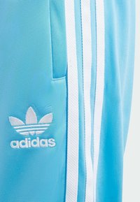 Joggers de color azul claro con rayas blancas clásicas de Adidas, que presentan un logo bordado en el lado izquierdo y un detalle de bolsillo lateral.