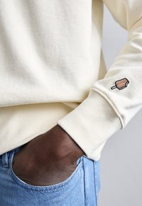 Mano di colore scuro in una tasca di jeans azzurri chiari, che indossa un maglione color crema con un piccolo ricamo di un ghiacciolo al cioccolato morso sulla manica.