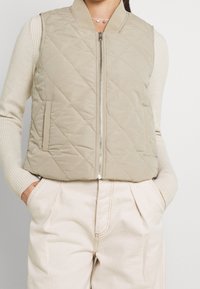 Jasno beżowa pikowana kamizelka z zapięciem na zamek, prążkowanym kołnierzem i kieszeniami bocznymi, w zestawieniu z kremowymi spodniami o wysokim stanie.