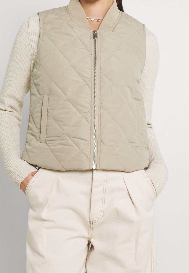 Jasno beżowa pikowana kamizelka z zapięciem na zamek, prążkowanym kołnierzem i kieszeniami bocznymi, w zestawieniu z kremowymi spodniami o wysokim stanie.