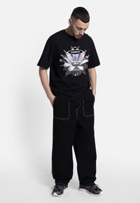T-shirt nera in cotone con una stampa grafica colorata sul davanti, abbinata a pantaloni neri con cuciture bianche e grandi tasche.
