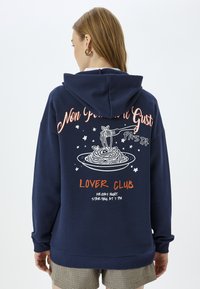 Granatowy hoodie z dużą grafiką makaronu na talerzu, z białym i pomarańczowym napisem. Materiał jest miękki, z luźnym krojem i kapturem.