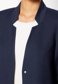 Cárdigan azul marino oscuro con cuello alto y cierre de botones a presión, confeccionado en una tela texturizada, combinado con una camiseta blanca acanalada.