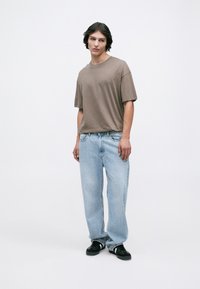 Young man with medium-length dark hair wearing a loose brown t-shirt, light blue jeans, and black sneakers standing against a white background.