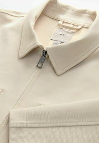 Veste beige zippée avec col. Fabriquée en tissu texturé, présentant des rayures diagonales et une fermeture éclair en métal. Étiquette visible avec le nom de la marque.