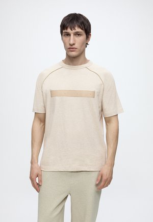 Man with short dark hair wears a beige short-sleeve shirt with "HUGO" text band across chest and light gray pants against white background.