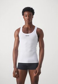 Fehér bordázott trikó kerek nyakkivágással, a mellkason egy kis fekete "VERSACE" logóval, fekete rövidnadrággal párosítva.