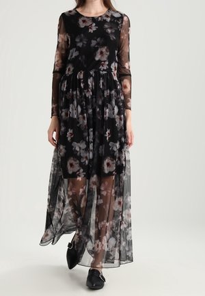 Czarna sukienka maxi w kwiaty z długimi, przezroczystymi rękawami, z talią podkreśloną paskiem i warstwową, przezroczystą spódnicą, ozdobiona różowymi i białymi kwiatowymi wzorami.