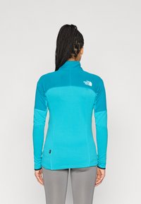 Ελαφρύ τοπ μακρυμάνικο σε φωτεινό τιρκουάζ με υφή στο σώμα, λείες μανσέτες και ψηλό γιακά. Διαθέτει μικρό λευκό λογότυπο στην πλάτη.